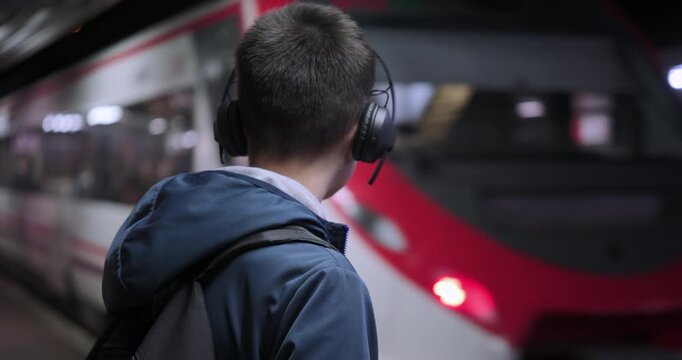 Rear view of a teenage boy with headphones and a backpack standing on a railway platform at night, watching the train arrive at the station.