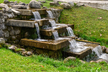 A charming, man-made tiered waterfall where clear water cascades over concrete steps, surrounded by rough stones and vibrant green mossy grass in a public garden in Razlog, Bulgaria. © Danica Chang
