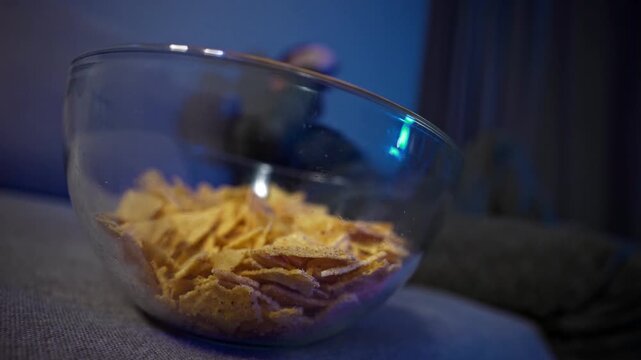 Late-night couch routine with TV and snacks. A man sits on a sofa in a dark room, illuminated by a TV screen. He eats chips, flips channels. Casual evening routine, screen addiction, passive rest.