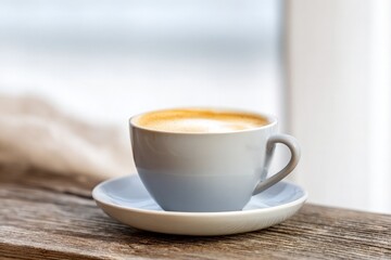 Warm cappuccino cup with creamy foam rests on rustic wooden table, creating peaceful scene near bright window.