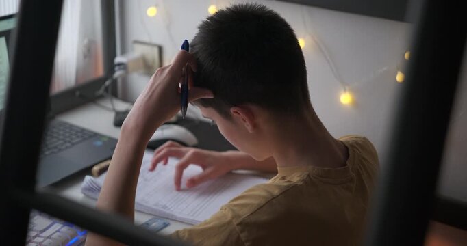 Pensive teenage boy holding a pen to his head while thinking about a difficult homework assignment, tapping fingers on a notebook in front of a laptop in a cozy room.