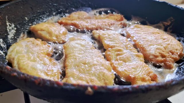 Mid-angle view of crispy golden-brown breaded fish fillets frying in bubbling hot oil inside a cast iron pan. The sizzling fat shows a delicious culinary preparation for a home-cooked dinner meal.