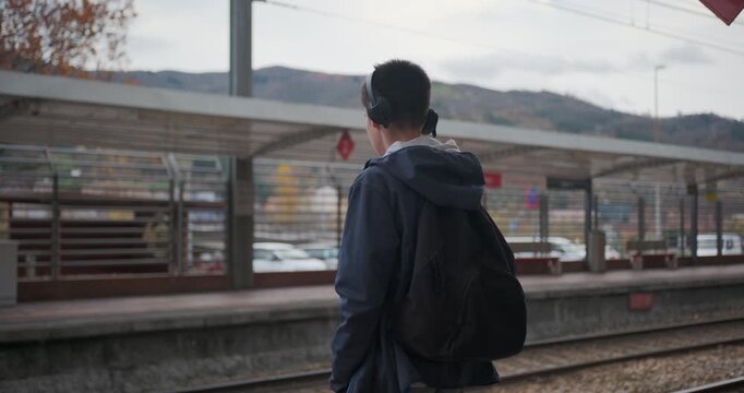 Rear view of a teenage boy with a backpack and headphones standing on a railway platform, looking around while waiting for a train on a cloudy day.