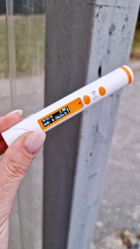 Close-up of a female hand holding a white pen-style radiation detector. The orange LCD screen displays a current dose rate reading, with the device held against a blurred grey concrete pillar outdoors
