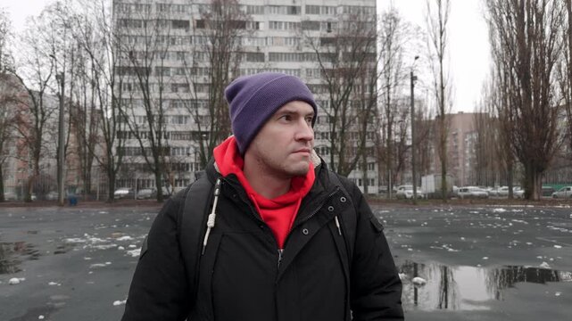 A man is standing in an open space surrounded by tall buildings. The ground is wet, and there is debris scattered around. He looks around while wearing a jacket and beanie.