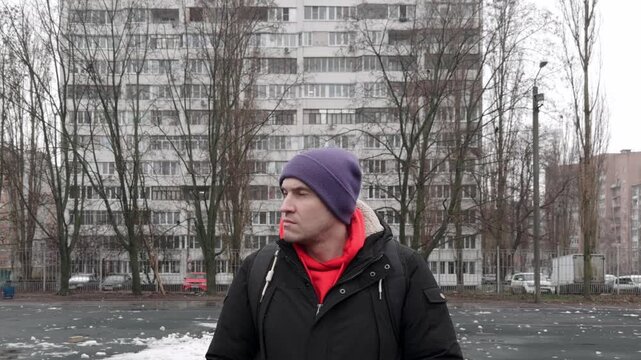 A man is standing in an open space surrounded by tall buildings. The ground is wet, and there is debris scattered around. He looks around while wearing a jacket and beanie.