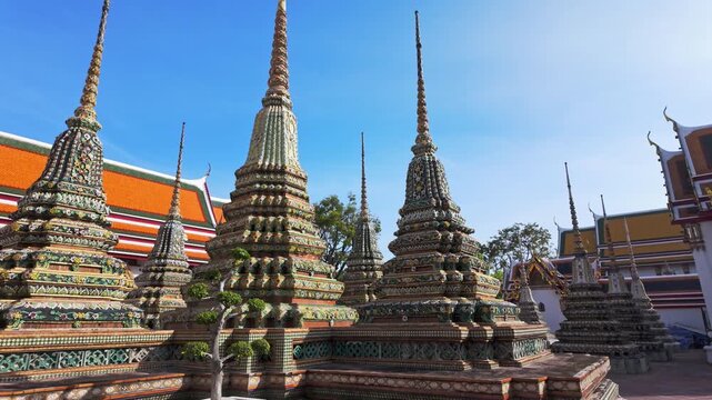 Exterior view of Wat Pho with richly decorated stupas covered in colorful floral ceramics and golden temple roofs shining under a clear blue sky on a bright sunny day in Bangkok.	
