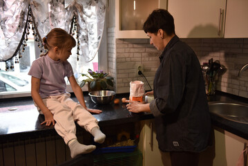 A woman prepares ingredients on the kitchen counter while a girl sits nearby, watching. Soft window light brightens the cozy kitchen as they get ready to bake together.