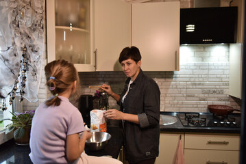 A woman prepares ingredients with a whisk while a girl sits on the counter beside a mixing bowl. Warm kitchen lighting highlights their shared cooking moment.