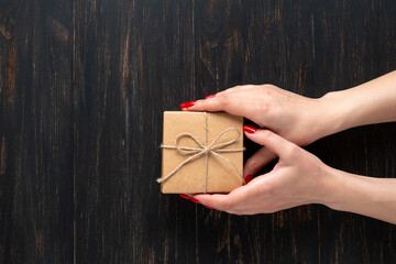 Female hands with red nails holding a small craft paper gift box on a dark wooden back