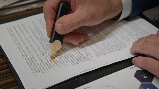 Close-up of hands of unrecognizable mature male attorney or businessman sitting at desk, analyzing contract, highlighting terms using marker, while working in office