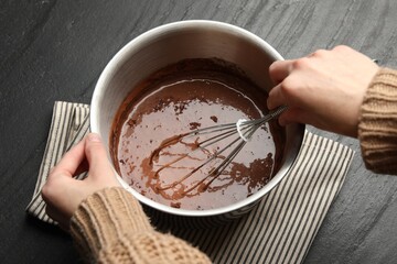 Woman mixing liquid chocolate dough with whisk at black table, above view