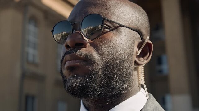 Close-up of face of young African American male security agent or guard, wearing suit and sunglasses, standing outdoors by government office, adjusting transparent earpiece while on duty