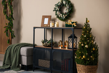 Shelving unit with Christmas decor, pouf and small spruce tree near beige wall indoors. Interior design