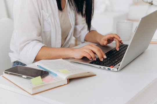 Hands of freelance woman typing on laptop keyboard over open notebook and phone, sticky notes and pen visible, closeup emphasizing fast efficient workflow, neat manicure and focused desk organization