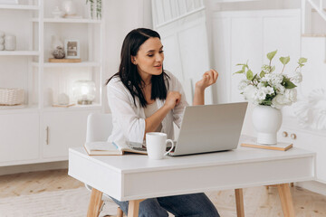 Caucasian freelancer woman smiling while reviewing moodboard on phone and sketching notes beside laptop, branding designer working in bright minimalist studio with floral decor and calm vibe