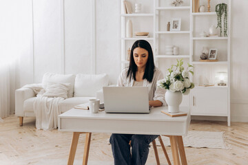 Caucasian freelancer woman focused on laptop spreadsheets while consulting phone notes, financial planner calculating budgets at tidy white desk with notebook and coffee, professional calm atmosphere
