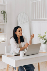 Top view of brunette woman typing text on a laptop on white office desk with table lamp, notebook, bottle of drinking whater and flowers in vase. Concept of freelancing, remote work, online learning