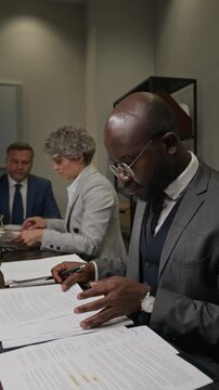 Medium vertical shot of busy African American male barrister in gray suit examining case files and evidence while preparing for court litigation at team meeting at law firm