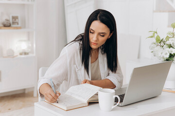 Young brunette freelancer woman sitting at desk using laptop writing notes while studying online, watching webinar, looking at pc screen learning web classes or remote working from home. Side view