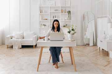Caucasian freelancer woman typing and referencing notes, creative copywriter editing drafts beside phone and notebook, white desk with floral accents and bright natural light, thoughtful mood