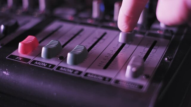 Extreme close-up of a finger pushing up the main volume fader on a professional audio mixer. A red effects fader is visible with dynamic colorful lighting changing from blue to purple.