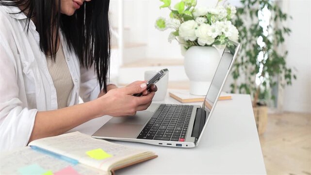 Closeup caucasian woman using smartphone and laptop, sticky notes and planner on desk, active social media manager or assistant multitasking in bright home office, focused hands and device interaction