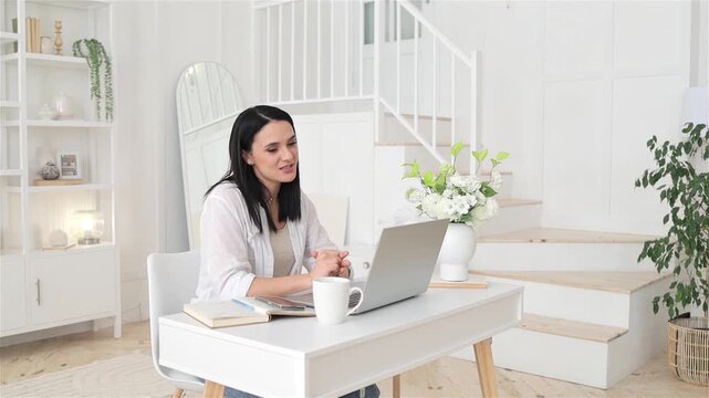 Caucasian woman at laptop, using expressive hand gestures in bright staircase studio, white desk with coffee and book, supportive teaching atmosphere, friendly online lesson or coaching session