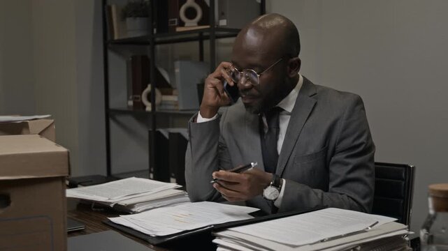 Medium shot of angry young African American male lawyer in gray suit sitting at desk alone at night in office, arguing on phone with colleague over mistakes in legal documents before deadline