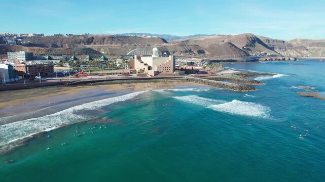 Scenic aerial footage of Las Palmas coastline with surfers riding waves on a bright sunny day. Gran Canaria