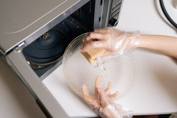 Close-up top view of female hands in disposable gloves cleaning microwave oven interior with yellow sponge, maintaining kitchen hygiene standards and ensuring household cleanliness.