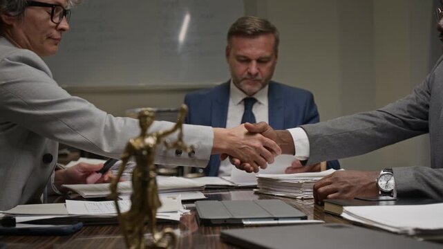 Medium shot of diverse male and female lawyers reviewing case documents at team meeting at legal firm, agreeing on contract clause and shaking hands, Lady Justice figurine on table in foreground