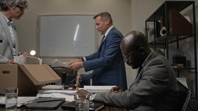 Medium shot of multiethnic attorneys working together at night in office, searching for precedents in archives, senior male partner finding information in case file and giving high fives to colleagues