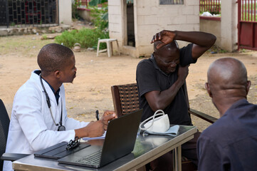 Young African male doctor listens attentively during an outdoor medical consultation as an adult male patient raises his arm and holds his shoulder in visible discomfort beside a table with medical eq