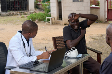 Young African male doctor observes and writes notes during an outdoor medical consultation as an adult male patient lifts his arm and reacts in visible discomfort while seated beside a table with medi