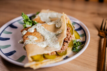 Close-up of a savory crepe with minced meat, pickles, greens, and creamy herb sauce, served on a patterned plate with soft warm lighting.