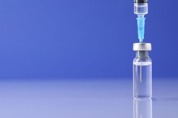 Filling syringe with vaccine from glass vial at light mirror surface against blue background, closeup. Space for text