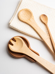 A collection of handcrafted wooden kitchen utensils, showing rustic natural textures. The set includes a spoon rest, serving utensils, and a white waffle weave dish towel. light and white background