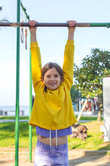 Fototapeta premium Smiling girl hanging on outdoor bar during active play, enjoying sunny weather and practicing strength and coordination in a park near the seaside, creating a bright and energetic childhood moment