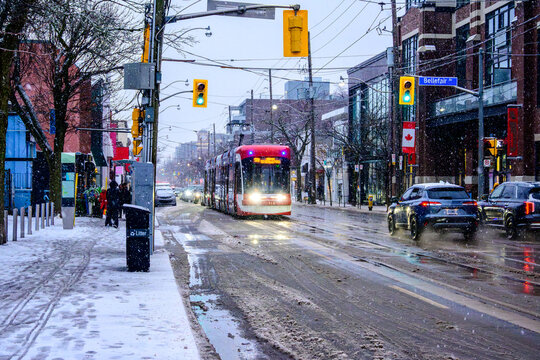Red Toronto streetcar at an intersection on Queen St East in the Beaches neighbourhood on a slushy December morning, surrounded by fresh snowfall, wet roads, passing traffic and traffic lights