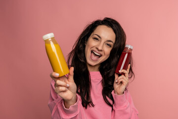 Cheerful beautiful young woman posing for the camera on the pink background with orange and tomato juice bottles in her hands