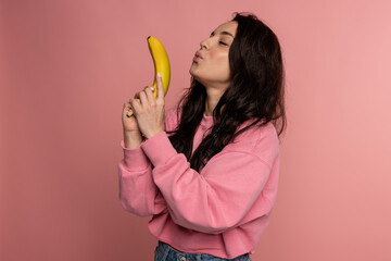 Young dark-haired brunette with a banana gun posing on the pink background during the studio photo shoot. Healthy food concept © Vlad