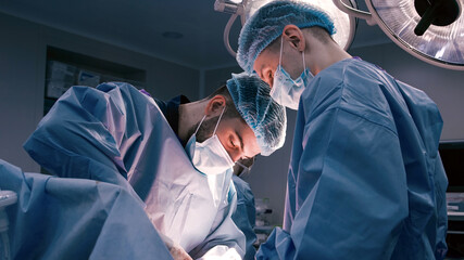 Two male surgeons in blue scrubs and surgical masks are focused on a patient during a procedure in a well-lit operating room, showcasing teamwork and precision in healthcare