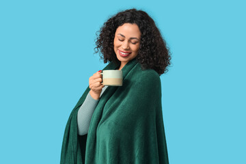 Beautiful young African-American woman with cozy blanket on blue background © Pixel-Shot
