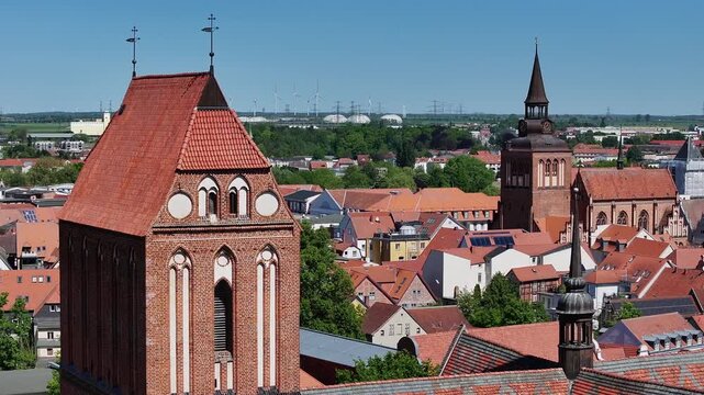 Telephoto drone flight to the left over G&uuml;strow, Mecklenburg-Western Pomerania, showing the cathedral and parish church, with industrial facilities, power lines, and a wind park in the background.