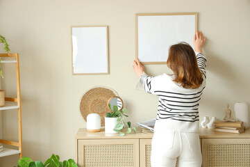 Young woman hanging blank frame on beige wall at home, back view