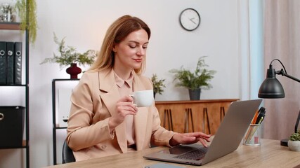 Middle-aged businesswoman at home office reads task list on laptop drinks coffee takes sip smiles stays focused. Freelancer girl at table enjoys cup gets energy plans steps types reply and saves file.