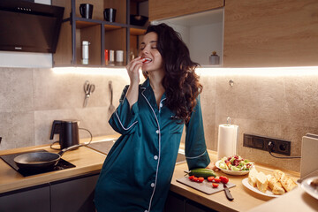 Happy young woman in silk robe eating tomato while standing in the kitchen and preparing dinner....