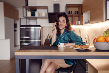 Smiling pretty brunette woman sitting at the bar counter at the kitchen in her flat, enjoying coffee, looking at camera. Morning routine, domestic life