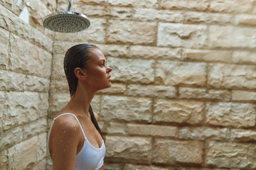 woman showering outdoors under rain showerhead with stone wall background, wet hair, relaxed expression, natural light, casual white top, summer wellness.